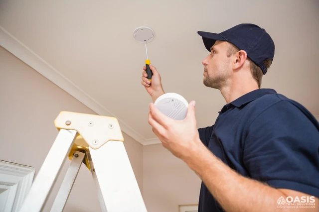 Technician installing a smoke detector on a ceiling using a ladder and screwdriver